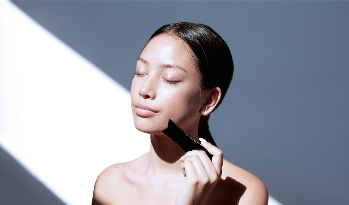 Asian model holding black obsidian gua sha stone tool. Facial massage tool from Traditional Chinese Medicine. 
